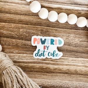 May include: A white sticker with the text "POWERED BY diet coke" in colorful, stylized lettering. The sticker is on a wooden surface.