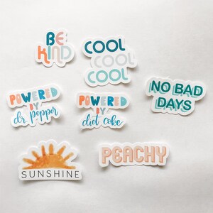 May include: A collection of colorful stickers with positive messages. The stickers include phrases like "Be Kind," "Cool," "Powered by Dr. Pepper," "Powered by Diet Coke," "No Bad Days," "Sunshine," and "Peachy."