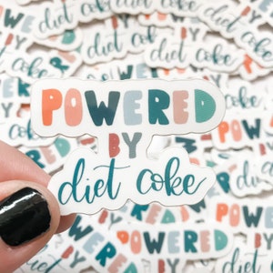 May include: A sticker with the text "POWERED BY diet coke" in a colorful, handwritten font. The sticker is white with a light blue, pink, and teal color scheme. The sticker is being held by a hand.