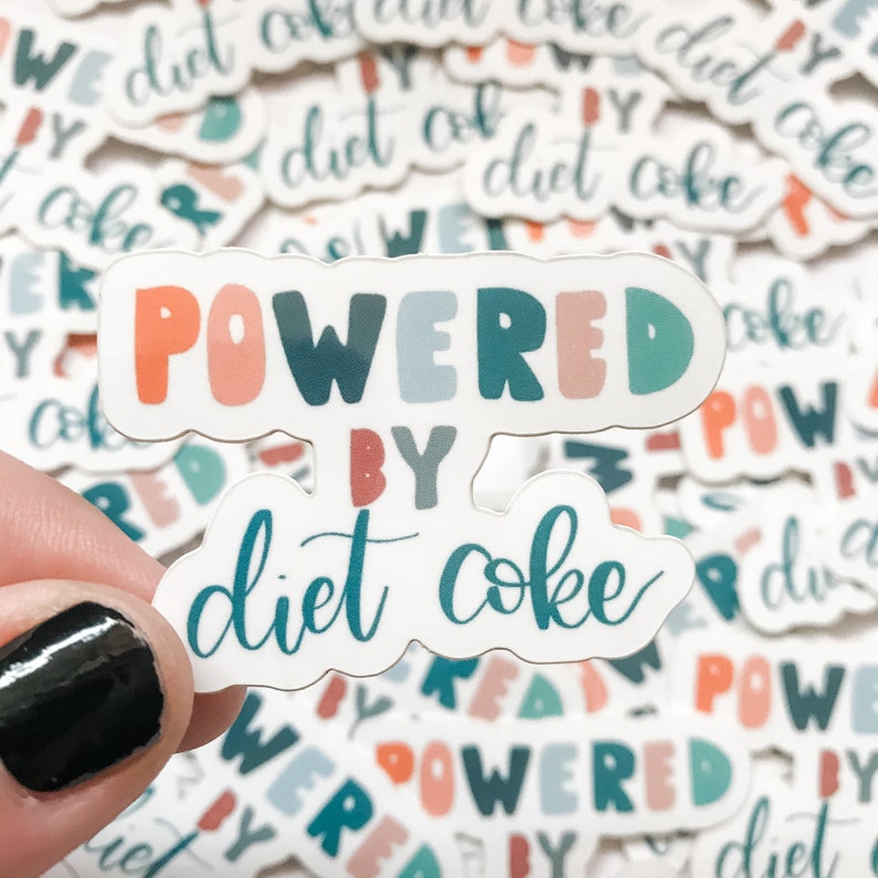 May include: A sticker with the text "POWERED BY diet coke" in a colorful, handwritten font. The sticker is white with a light blue, pink, and teal color scheme. The sticker is being held by a hand.