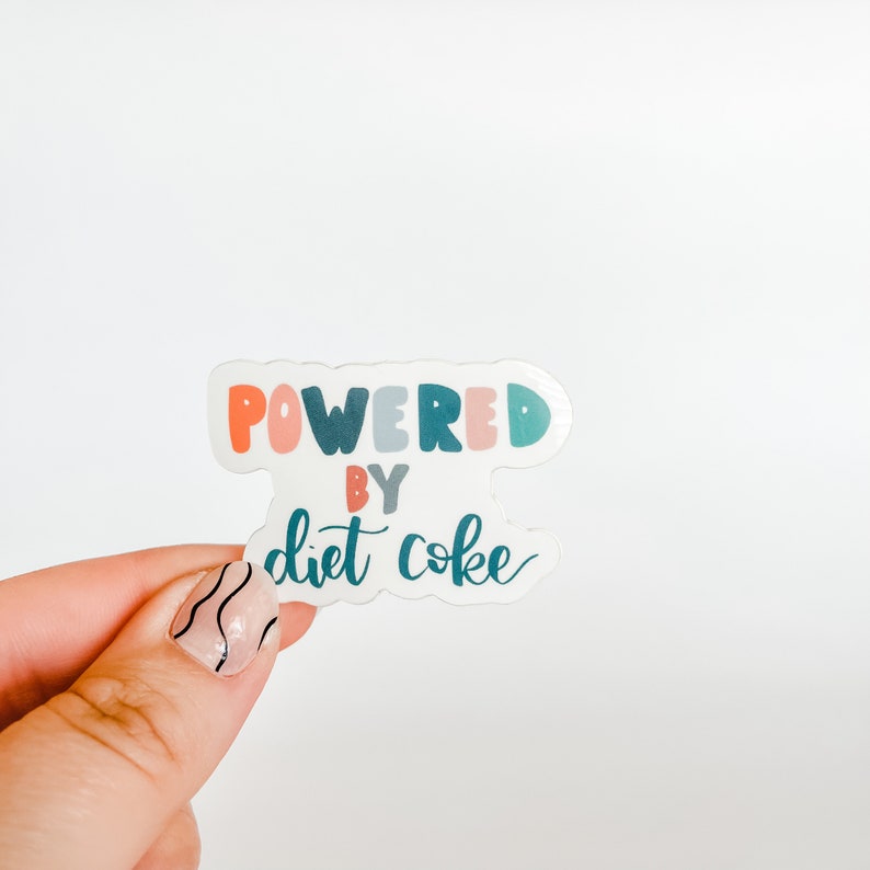 May include: A white sticker with the text "Powered by Diet Coke" in colorful letters. The sticker is held in a hand with black nail polish.