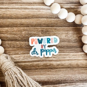 May include: A sticker with the text "POWERED BY dr. pepper" in a colorful, playful font. The sticker has a white border and is set against a wooden background. Decorative wooden beads and a tassel are also visible.