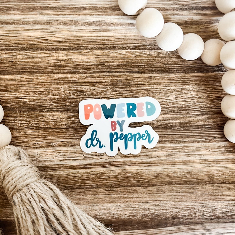 May include: A sticker with the text "POWERED BY dr. pepper" in a colorful, playful font. The sticker has a white border and is set against a wooden background. Decorative wooden beads and a tassel are also visible.