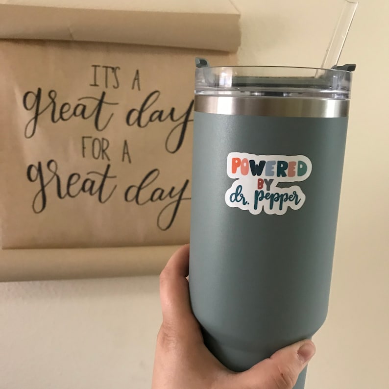 May include: A large, teal-colored tumbler with a clear straw and a silver rim. A sticker on the tumbler reads "Powered by Dr. Pepper" in colorful text. In the background, a scroll displays the phrase "It's a great day for a great day."