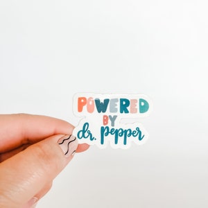 May include: A colorful sticker with the text "POWERED BY dr. pepper" in a playful font. The sticker features orange, pink, teal, and blue lettering. The sticker is held against a white background.