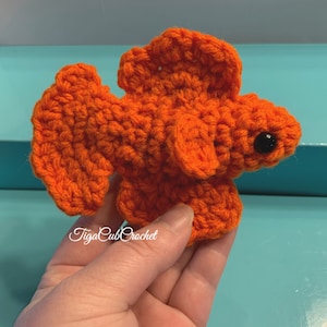 May include: Orange crocheted betta fish with black eyes. The fish is being held in a hand.