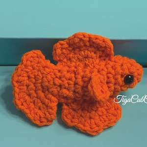 May include: Orange crocheted betta fish with black eyes. The fish is on a blue surface. The text "TigaCubCrochet" is in the lower right corner of the image.