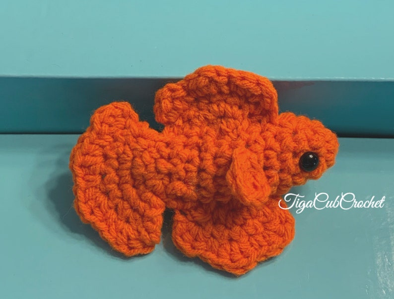 May include: Orange crocheted betta fish with black eyes. The fish is on a blue surface. The text "TigaCubCrochet" is in the lower right corner of the image.