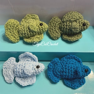 May include: Four crocheted betta fish, two green and two blue. The green fish are on a light blue surface and the blue fish are on a turquoise surface. The fish are all about 3 inches long.
