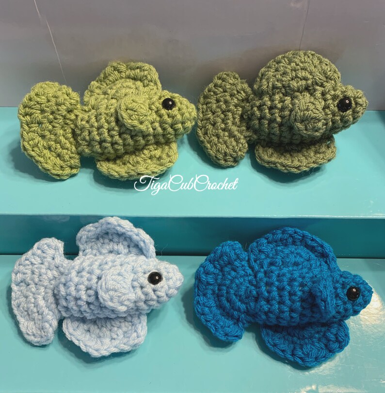 May include: Four crocheted betta fish, two green and two blue. The green fish are on a light blue surface and the blue fish are on a turquoise surface. The fish are all about 3 inches long.