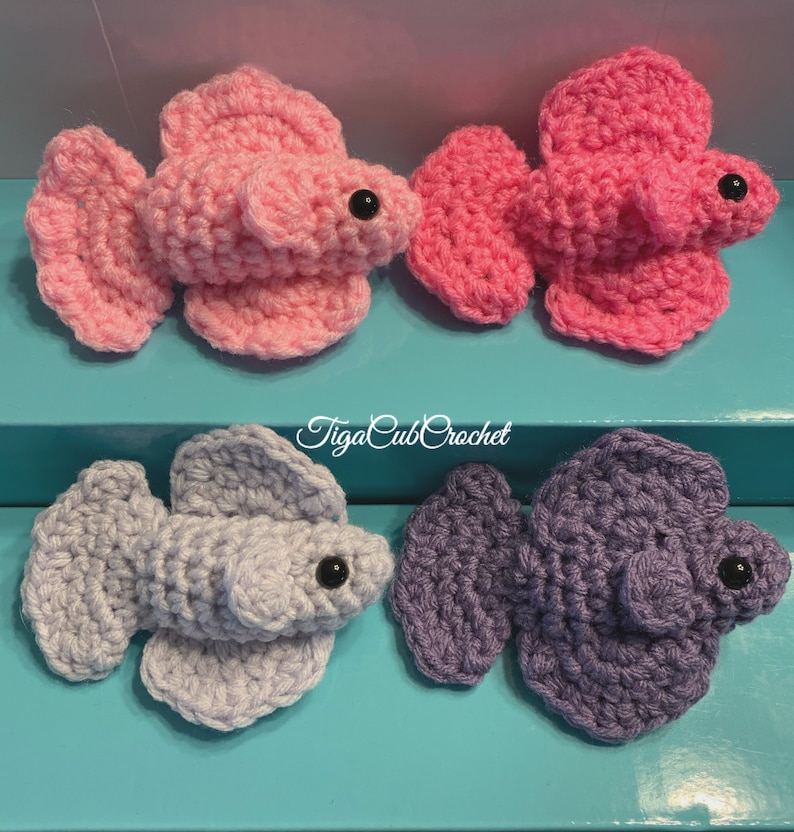 May include: Four crocheted betta fish, two pink, one light purple, and one gray. The fish are all in a similar pose with their fins spread out. The fish are all about 3 inches long.