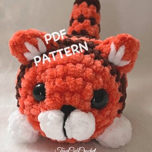 Könnte beinhalten: Ein gehäkeltes Tiger-Amigurumi-Spielzeug mit orangefarbenem und braunem Fell, weißen Pfoten und schwarzen Augen. Das Spielzeug liegt auf dem Bauch mit eingerolltem Schwanz. Der Text "PDF PATTERN" ist in Weiß auf rotem Hintergrund geschrieben.