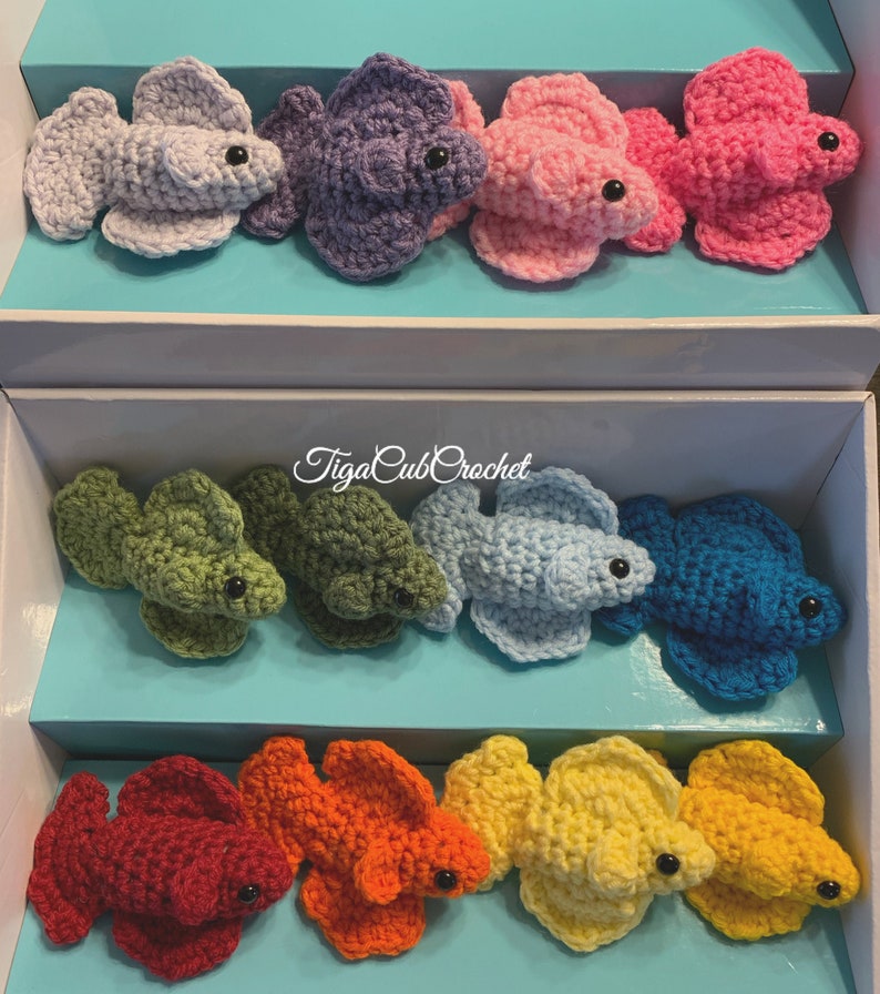 May include: A collection of ten crocheted betta fish in various colors, including purple, pink, green, blue, red, orange, and yellow. The fish are arranged in two rows of five, with the text "TigaCubbCrochet" printed on the box.