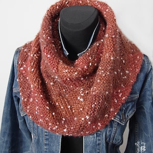 May include: A hand-knitted cowl scarf in shades of rust and brown, speckled with white flecks. The chunky knit scarf is displayed on a black mannequin head over a denim jacket, showcasing its texture and colour variations. A cosy accessory.