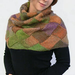 May include: A colourful knitted cowl with a geometric pattern in shades of green, orange, brown, and purple. The cowl is worn by a person with a black shirt.