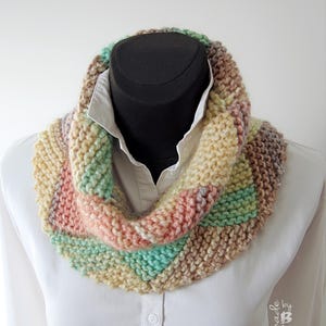May include: A knitted cowl scarf in pastel shades, displayed on a mannequin. The scarf features a mix of soft colours including pink, green, beige, and brown. The cowl is thick and appears hand-knitted, with a ribbed texture. The mannequin is wearing a white collared shirt.