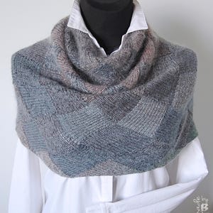 May include: A knitted cowl scarf in shades of grey, blue, and pink, featuring a geometric design. The scarf is displayed on a black mannequin head over a white collared shirt. The handmade scarf has a textured appearance.