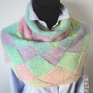 May include: A hand-knitted cowl scarf in pastel shades. The scarf has a patchwork design with squares of pink, green, lavender, and yellow. The soft, chunky knit scarf is displayed on a mannequin with a light blue collared shirt.