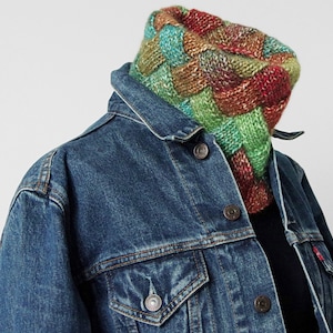 May include: A colourful knitted neck warmer with a braided pattern in shades of green, red and brown. The neck warmer is worn over a denim jacket.