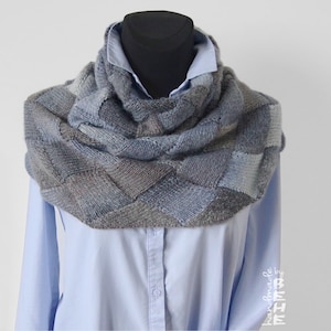 May include: A grey and blue knitted infinity scarf with a geometric pattern. The scarf is displayed on a mannequin wearing a light blue shirt.