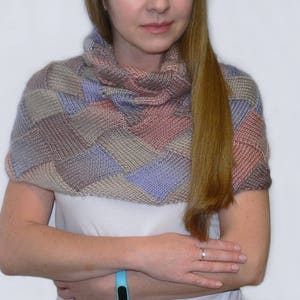 May include: A knitted cowl with a patchwork design in shades of pink, purple, and brown. The cowl is worn by a person with long brown hair.