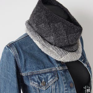 May include: A knitted cowl scarf in shades of grey and charcoal. The scarf is displayed on a mannequin, worn over a denim jacket and a black top. The cowl has a gradient effect, changing from light grey to charcoal.