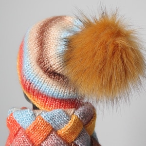 May include: A colourful knitted hat with a large orange fur pom pom. The hat is made with a striped pattern of orange, brown, blue and white yarn.