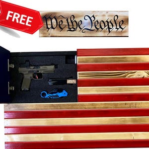 May include: A wooden American flag with red, white, and blue stripes. The flag has a hidden compartment that opens to reveal a gun and a small blue tool. The flag also has a wooden sign above it that says "We the People".