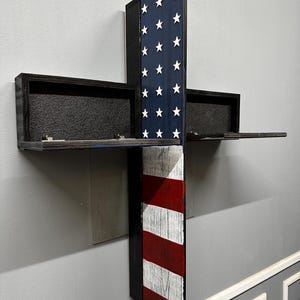 May include: A wooden wall-mounted shelf in the shape of a cross, painted with the American flag design. The top section features a blue background with white stars, while the bottom has red and white stripes. The shelf has two black storage compartments.