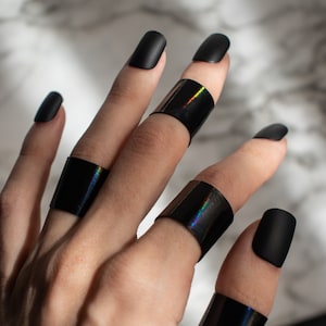 May include: A hand wearing five black nail polish rings with iridescent accents. The rings are worn on the index, middle, ring, and pinky fingers.