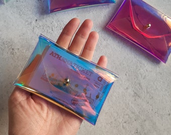 May include: A clear iridescent envelope-style wallet with a silver button closure. The wallet is holding a ticket with the text "ATHENATICKET" printed on it.