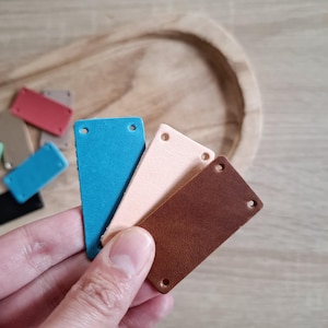 May include: Four leather labels in different colours: teal, light pink, brown, and red. Each label has two holes punched in the top.