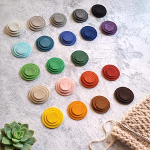 以下が含まれることがあります： 青、緑、ピンク、茶色など、さまざまな色合いのカラフルな重ねられたフェルトサークル。 サークルは、小さな多肉植物と編み針のある編み物と一緒に、明るい灰色の表面に配置されています。