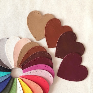 Op de afbeelding: Hartvormige leren stukken in verschillende kleuren, waaronder bruin, beige en bordeaux. Elk stuk heeft voorgeponste gaten voor het naaien. Een kleurenpalet van leren stalen is in een halve cirkel gerangschikt.