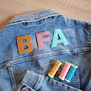 Puede incluir: Chaqueta vaquera azul con las letras "BFA" cosidas en la espalda en marrón, rosa y turquesa. Tres carretes de hilo de colores a juego se colocan debajo, lo que sugiere un proyecto de bricolaje o personalización.