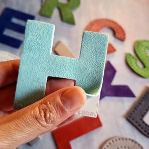 May include: A light blue suede letter 'H' is held in a hand, with other colourful felt letters in the background. The letter has a peel-off adhesive backing. The word 'WORDS' is partially visible on the backing.