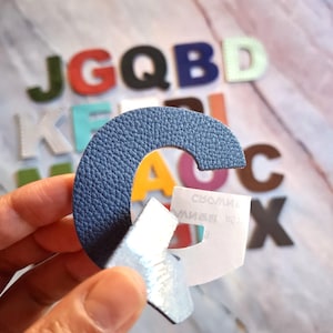 Può includere: Primo piano di una lettera "G" in pelle blu con una superficie strutturata, tenuta in mano. Altre lettere colorate sono sullo sfondo. La lettera "G" ha un supporto di carta.