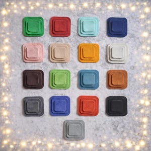 Pode incluir: Uma variedade de bases para copos quadradas de feltro coloridas em várias tonalidades, incluindo verde, vermelho, azul e laranja. Cada base para copo tem um design em camadas, criando um efeito tridimensional. As bases para copos estão dispostas sobre uma superfície cinza texturizada.