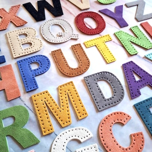 May include: A collection of colourful, stitched leather letters in various fonts. The letters are in shades of blue, yellow, green, orange, red, purple, white, black, and grey. Each letter has a unique colour and is arranged on a light-coloured surface.