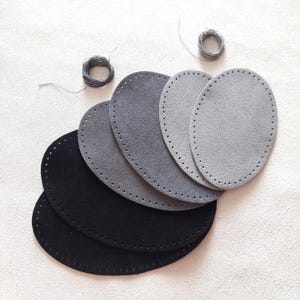 May include: A selection of oval suede leather bases in black and various shades of grey, each with pre-punched holes for stitching. Two small spools of grey thread are also visible. These are likely for crafting, such as bag making or similar projects.