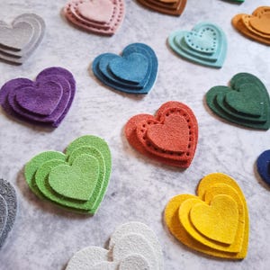 Könnte beinhalten: Eine Auswahl an mehrlagigen herzförmigen Filzverzierungen in verschiedenen Farben, darunter Lila, Blau, Grün und Gelb. Einige Herzen haben gestickte Details. Der Filz hat eine strukturierte Oberfläche.