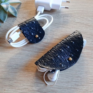 May include: Two black leather cord organizers with gold accents. One organizer is holding a white charging cable and the other is holding white earbuds.
