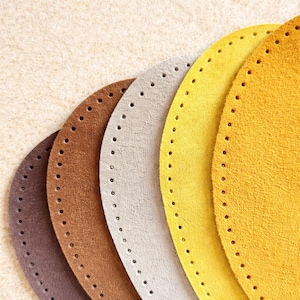 May include: Close-up of several oval-shaped leather bases in shades of brown, tan, and yellow. Each base has a series of evenly spaced holes around the edge, suggesting they are for crafting or sewing projects. The textures vary.