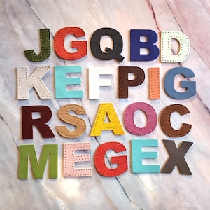 Peut inclure: Un ensemble de lettres colorées, en simili cuir, disposées sur une surface en marbre. Les lettres sont de différentes couleurs, dont le vert, le rouge, le noir, le bleu, le blanc, le marron et le rose. Chaque lettre a une bordure cousue.