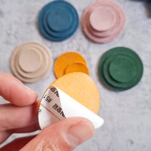 May include: A selection of colourful, round, adhesive pads in shades of blue, pink, green, beige, and orange. One pad has its backing partially peeled off, revealing the adhesive. The pads have a layered design.