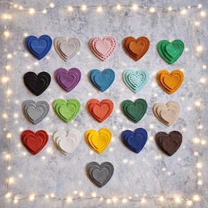 Op de afbeelding: Een verzameling gelaagde vilten hartdecoraties in verschillende kleuren, waaronder blauw, roze, groen en oranje. Sommige harten hebben decoratieve stiksels. De harten zijn gerangschikt op een getextureerd grijs oppervlak, met lichtslingers op de achtergrond.