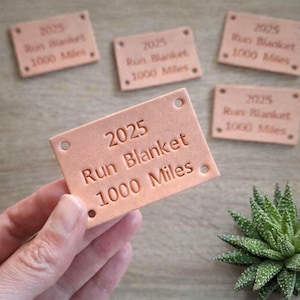 May include: A collection of rectangular, light brown leather tags. Each tag has the text "2025 Run Blanket 1000 Miles" embossed on it. The tags have two holes at the top and a single dot at the bottom.