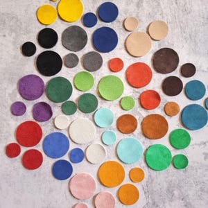 May include: A selection of multi-coloured circular leather pieces in different sizes, including yellow, black, blue, red, green, and pink. The pieces are arranged on a textured, light grey surface, creating a vibrant and artistic display.