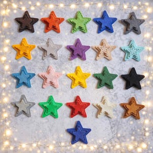 May include: A selection of multi-coloured felt stars in shades such as brown, orange, green, blue, yellow, grey, purple, pink, and black. Each star has a layered design, creating a 3D effect. The stars are arranged on a light grey surface.