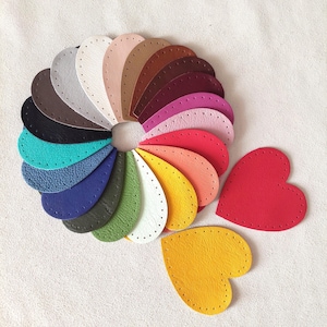 Op de afbeelding: Een verzameling kleurrijke leren hartvormige en druppelvormige uitsparingen, gerangschikt in een cirkelvormig patroon. De kleuren omvatten tinten rood, geel, groen, blauw en bruin. Elk stuk heeft voorgeponste gaten om te naaien.